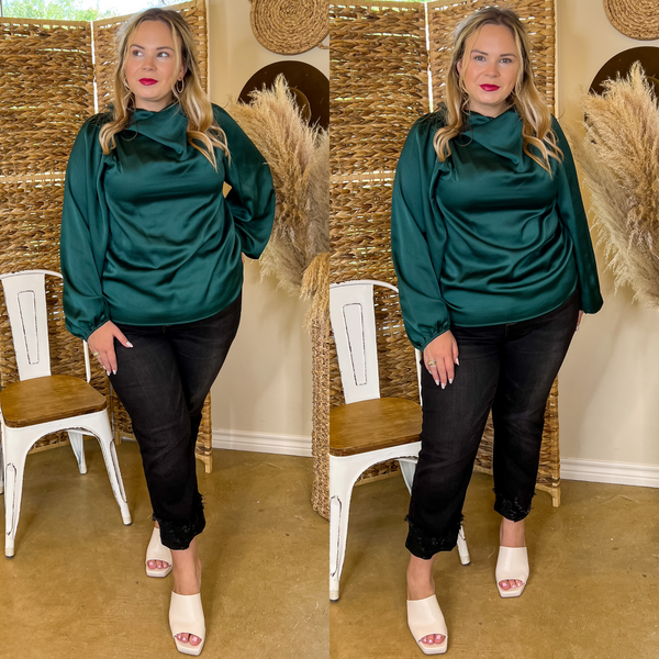 Model is wearing an emerald green long sleeve cowl neck top. Model has it paired with black jeans, white heels, and gold jewelry.