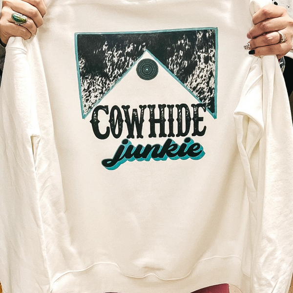 A long sleeve white sweatshirt with a cowhide and turquoise graphic that says "Cowhide Junkie."