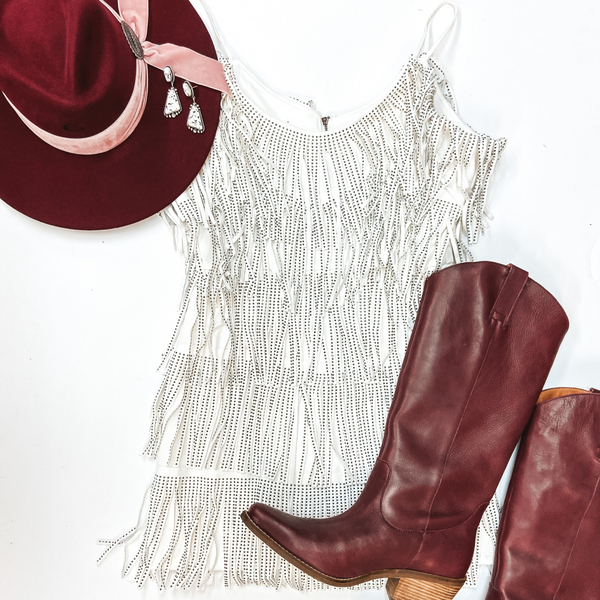 A white fringe crystal dress with spaghetti straps. Pictured on white background with burgundy boots and a burgundy hat.