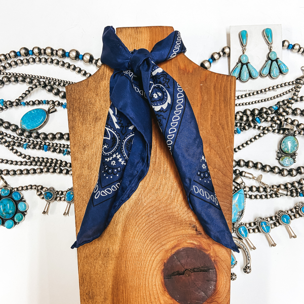 A navy blue silky wild rag with white bandana print tied around a wooden display. Pictured on white background with silver and turquoise jewelry.