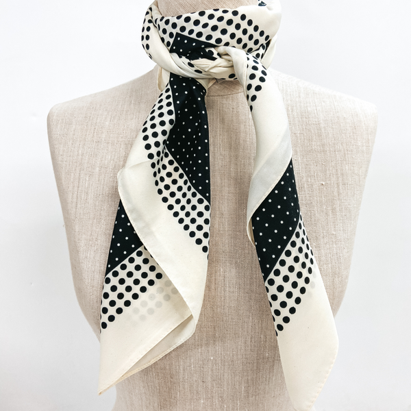 A black and ivory polka dot scarf tied around the neck of a mannequin. Pictured on white background.