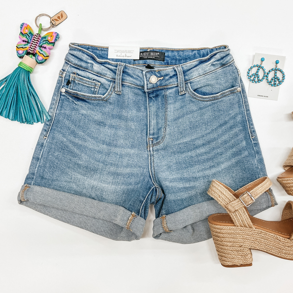 A pair of cuffed light wash shorts pictured on a white background with a beaded butterfly keychain, espadrille shoes, and peace sign earrings.