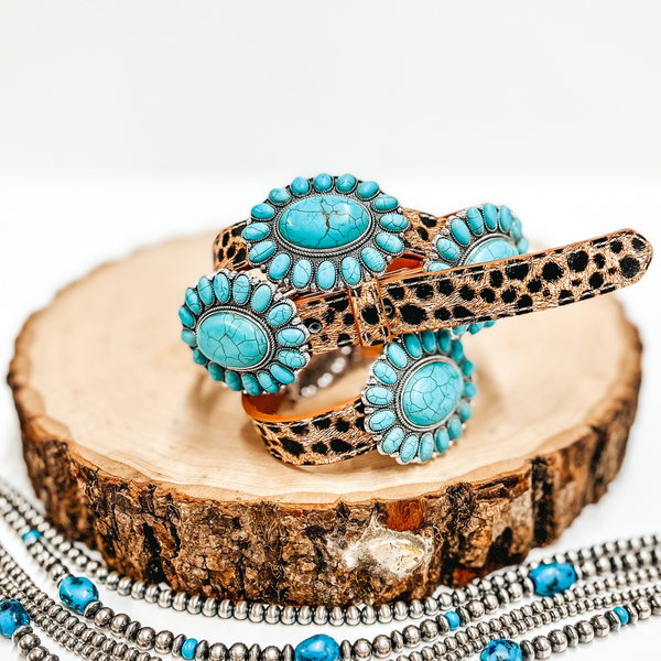 A leopard print belt with faux turquoise cluster conchos pictured on wooden display with Navajo pearls on a white background.