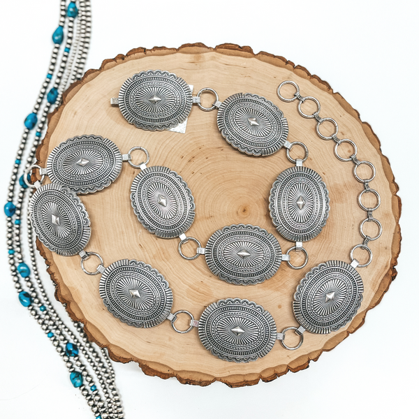 An oval silver concho belt placed on a wooden display. Pictured on white background with Navajo pearls.