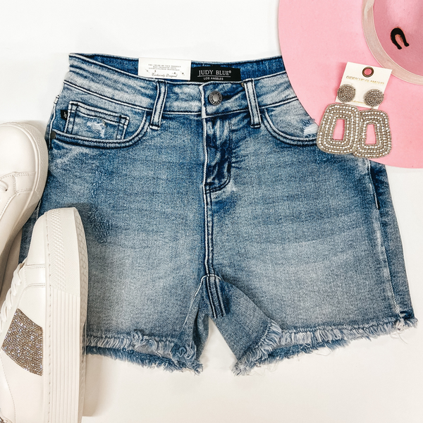 A pair of light wash shorts with a cut off hemline. Pictured with white crystal sneakers, crystal earrings, and a pink hat.