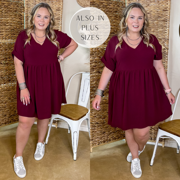 Model is wearing a maroon v neck babydoll dress with short sleeves. Model has it paired with white sneakers and silver jewelry.