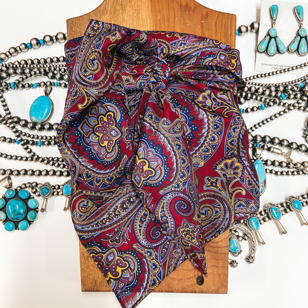 A calico paisley wild rag with maroon and blue print. Pictured around a wooden display on a white background with silver and turquoise jewelry.
