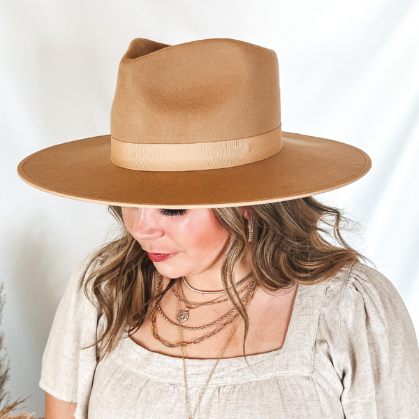 Model is wearing a light brown hat with a light brown ribbon band. Model has it paired with a beige top and gold jewelry.