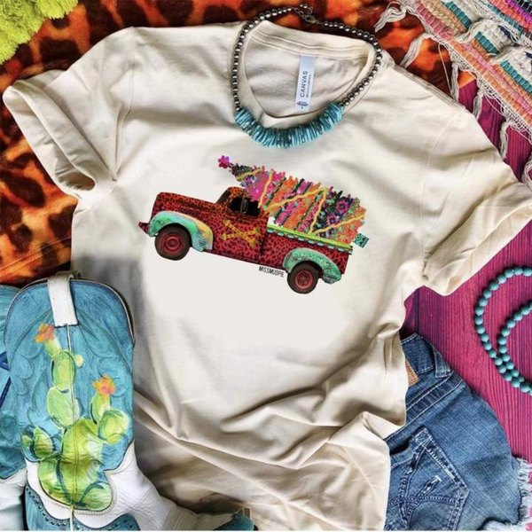 Model is wearing a crew neck tee shirt with short sleeves and a graphic of a leopard pick up truck, hauling a serape christmas tree. Pictured with a turquoise necklace and cowgirl boots