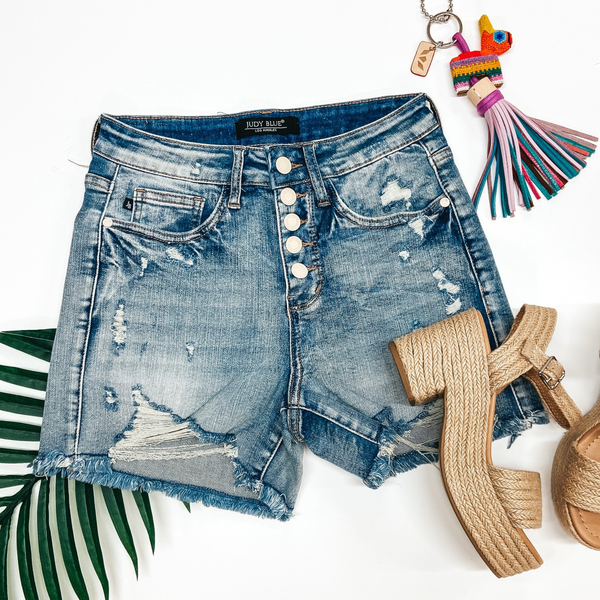 A pair of distressed denim shorts with a button fly. Pictured on a white background with heeled sandals and a palm leaf.