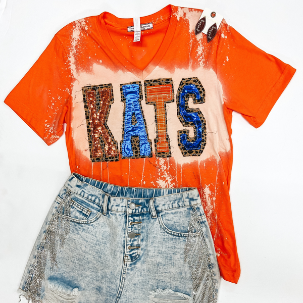 An orange tee shirt with a v neckline, short sleeves, and bleach splatter on the front. The tee shirt has patches that spell out "KATS" in orange and blue lettering with a leopard print trim.  This tee is pictured with beaded football earrings and denim shorts with crystal fringe.