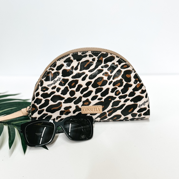 A leopard print dome cosmetic bag with leather zipper. Pictured on white background with a palm leaf and black sunglasses.