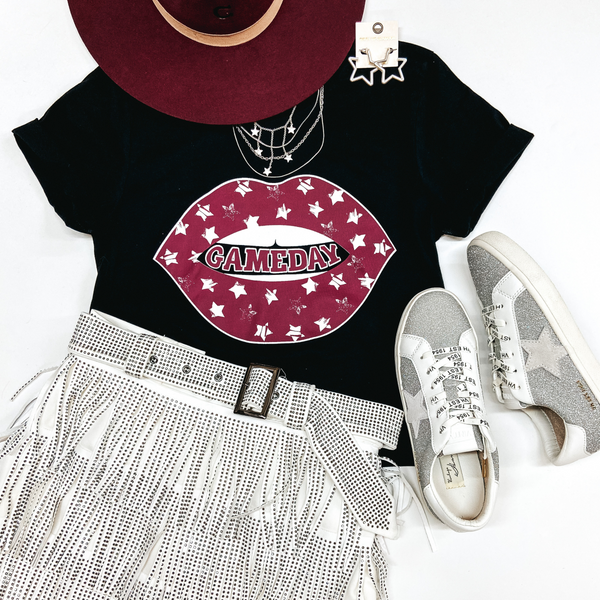 A black graphic tee with short sleeves and a maroon image of lips with stars that says "gameday" in between the lips. This tee shirt is pictured on a white background with a maroon hat, silver jewelry, a white crystal fringe skirt, and silver glitter sneakers.