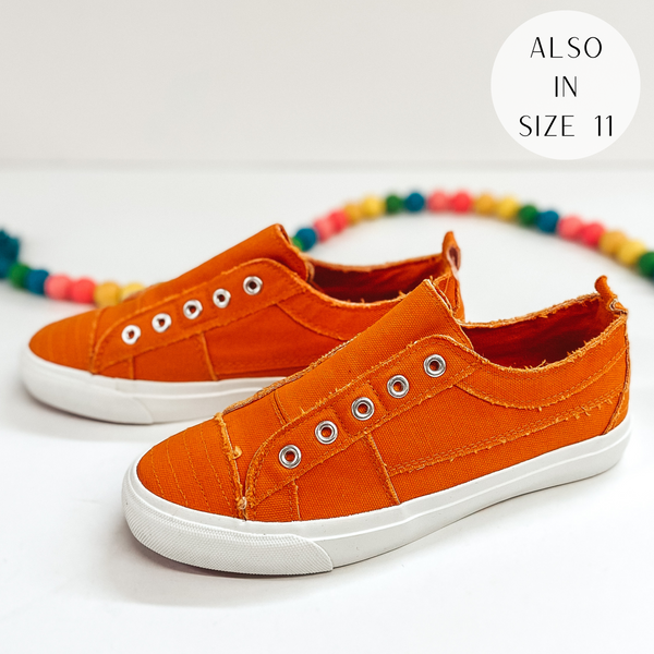 A pair of orange sneakers that do not have laces. Pictured on white background with colorful beads.
