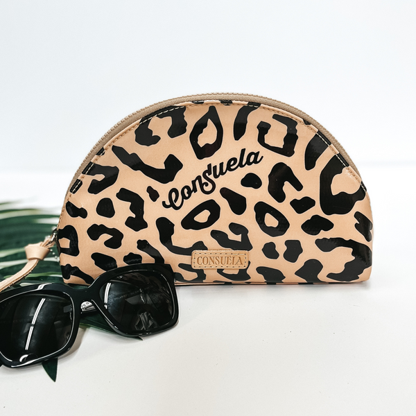 A dome shape leopard print cosmetic bag with a leather zipper. Pictured on white background with black sunglasses and a palm leaf.