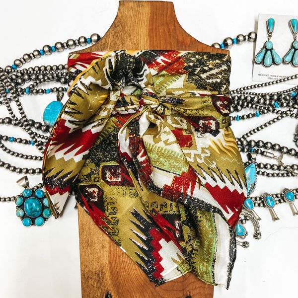 An Aztec print scarf with red, green, and white colors. Scarf is tied around a wooden display with turquoise and silver jewelry.
