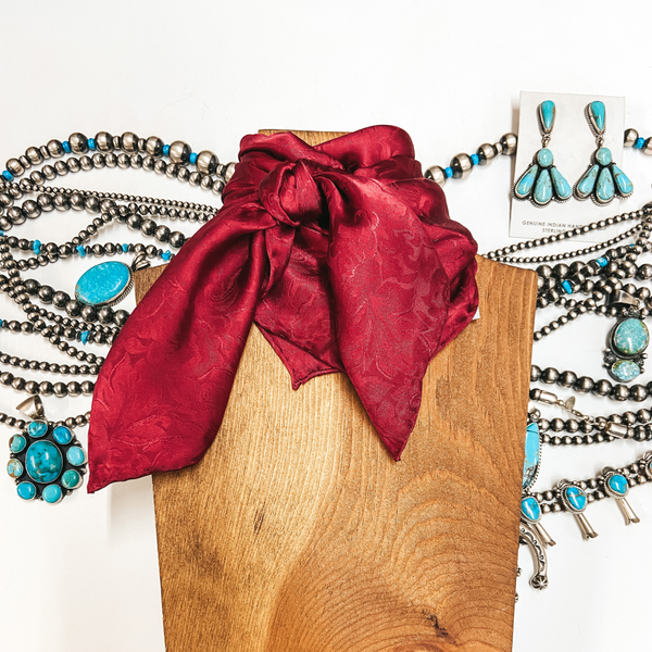 A red silky scarf with a jacquard print. Scarf is wrapped around wooden display. Pictured on white background with silver and turquoise jewelry.