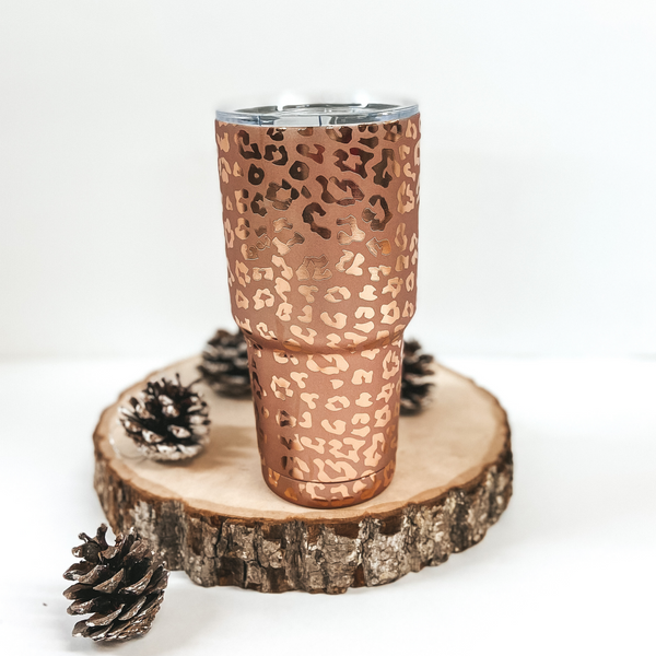 A matte rose gold tumbler with shiney rose gold leopard spots. Pictured on white background with pinecones and log.