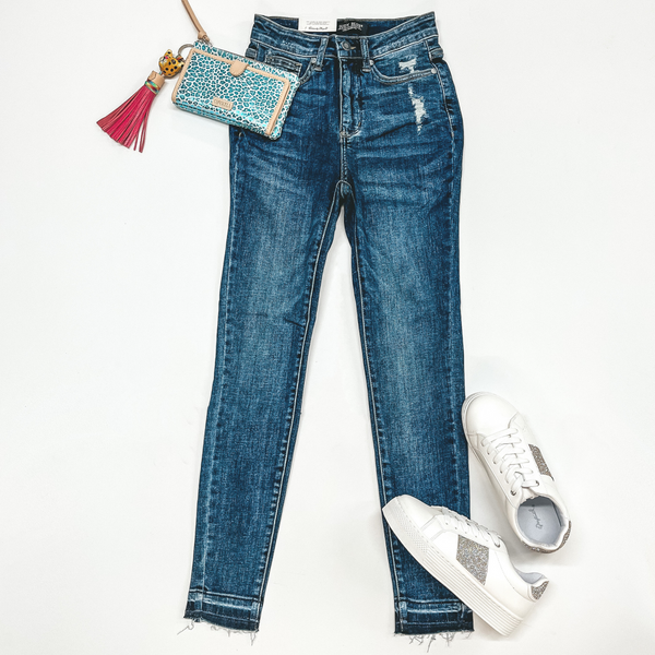A Pair of dark wash skinny jeans with a raw hem. Pictured on a white background with white sneakers, a blue leopard print wallet, and keychain.