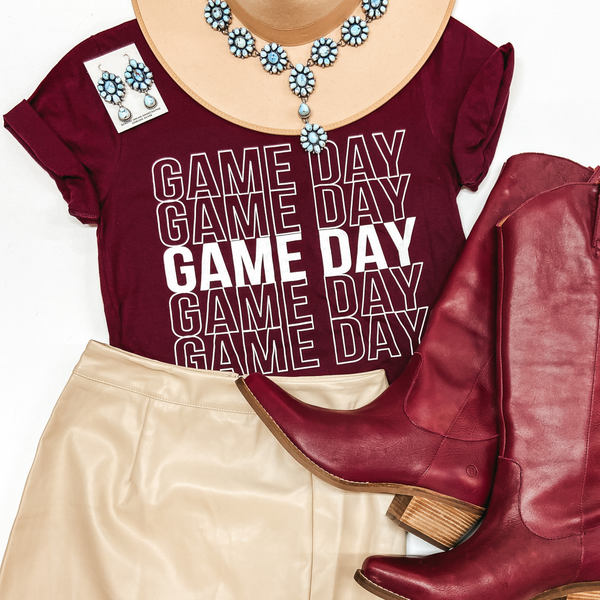 A maroon short sleeve graphic tee that says "Gameday" 5 times underneath each other. This tee is pictured with a tan hat, tan skirt, maroon boots, and turquoise jewelry.