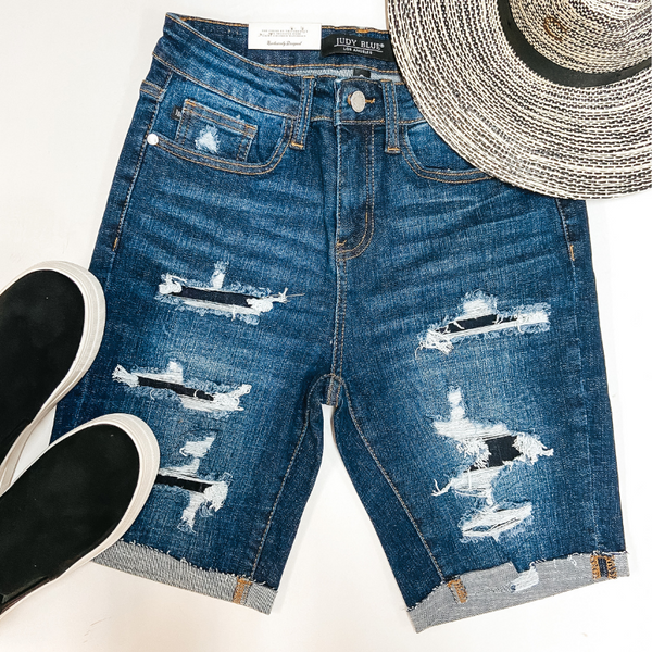 A pair of distressed bermuda shorts with patches. Pictured on white background with a straw hat and black sneakers.