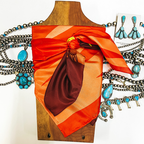 An orange mix silky scarf tied around a wooden display. Pictured on white background with turquoise and sterling silver necklace.