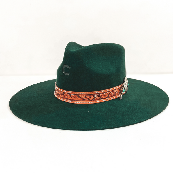 A dark green felt hat with a pinched front and flat brim. Hat has a brown leather band with a silver concho and silver removeable arrow pendant. Pictured on white background.