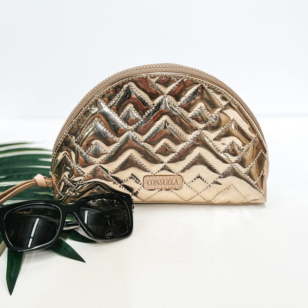 A gold dome cosmetic bag with a leather zipper. Pictured on white background with black sunglasses and a palm leaf.
