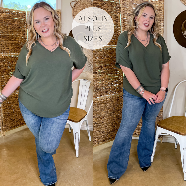 Model is wearing an olive green top with short sleeves and a v neck. Model has it paired with flare jeans, black boots, and silver jewelry.