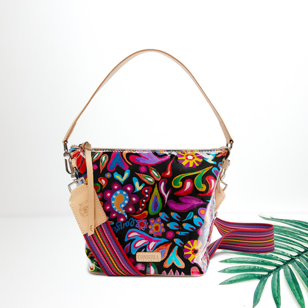 A black crossbody bag with a multi color paisley print. This bag is pictured on a white background with a palm leaf.