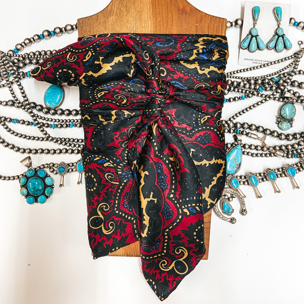 A red, black, gold, and blue paisley silky scarf tied around a wooden display. Pictured on white background with silver and turquoise jewelry.