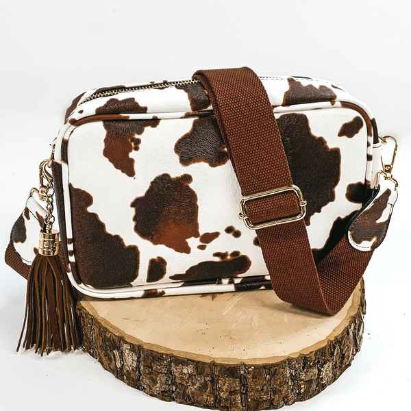 White and brown cow print rectangle purse with a dark brown adjustable strap and dark brown decorative tassel. This purse also includes gold accents. This purse is pictured sitting on a piece of wood on a white background.