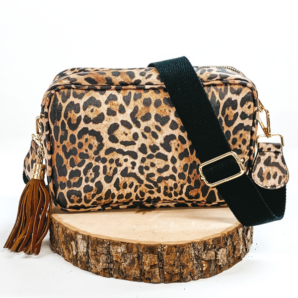Leopard print rectangle purse with a black adjustable strap and brown decorative tassel. This purse also includes gold accents. This purse is pictured sitting on a piece of wood on a white background.