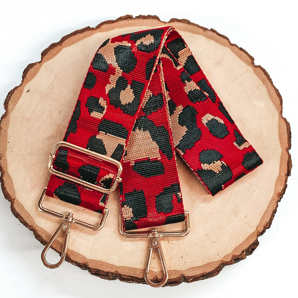 Shiny red purse strap with a tan and black leopard print. This purse strap includes gold accents. This purse strap is pictured on a piece of wood on a white background. 
