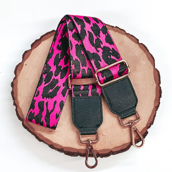 Fuchsia purse strap with a black leopard print. This purse strap includes gold accents and black pieces at each end. This purse strap is pictured on a piece of wood on a white background. 