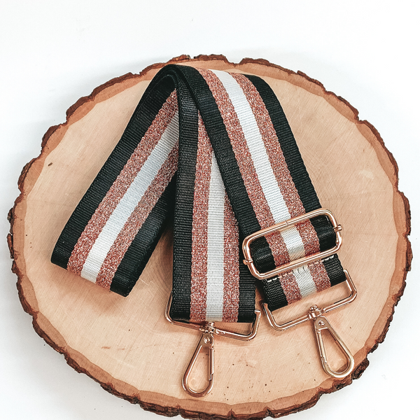 Black, sparkly rose gold, and white striped purse strip. This purse strap includes gold accents. This purse strap is pictured on a piece of wood on a white background. 
