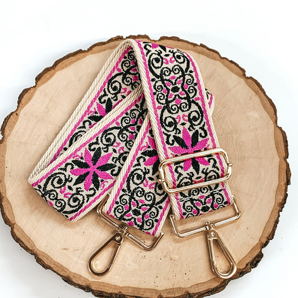Beige canvas purse strap with black and fuchsia embroidered design. This purse strap includes gold accents. This purse strap is pictured on a piece of wood on a white background. 