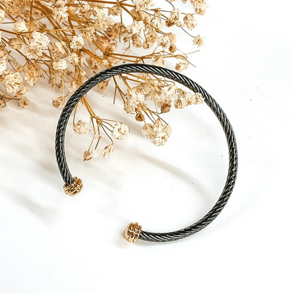 Charcoal cable bangle with gold cabochon ends. This bracelet is pictured on a white background with baby's breath on the top left corner. 