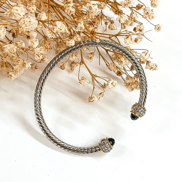Silver cable bangle with black crystal and silver and gold accented cabochon ends. This bracelet is pictured on a white background with baby's breath on the top left corner. 
