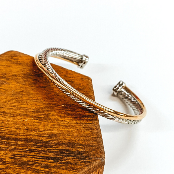 Silver cable band twisted around with a plain gold band bangle. This bracelet has silver end caps. This bracelet is pictured laying partially on top of a brown block.