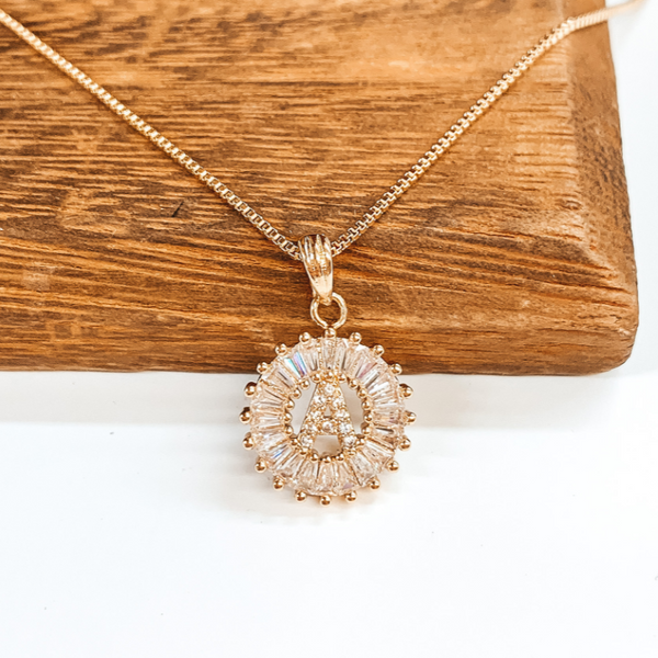 Gold chained necklace with crystal, open, circle pendant with a center crystal "A". This necklace is pictured laying on a brown block on a white background.