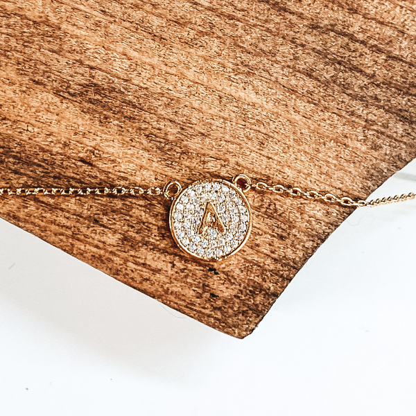 Thin gold chained necklace with circle, gold pendant. The pendant is covered with clear crystals and has a center gold "A". This necklace is pictured laying on a brown block on a white background. 