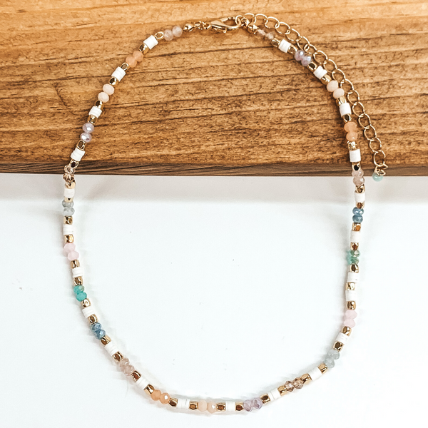 This necklace has segments of pastel crystal beads and white rubber beads with gold bead spacers. This necklace includes the pastel colors of blue, purple, green, ivory, and beige. This necklace is pictured on a laying halfway on a brown block on a white background. 