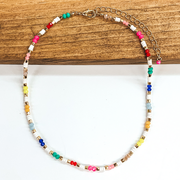 This necklace has segments of bright crystal beads and white rubber beads with gold bead spacers. This necklace includes the bright colors of blue, yellow, green, red, pink, and beige. This necklace is pictured on a laying halfway on a brown block on a white background.