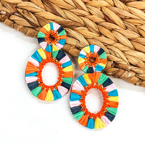 Circle, open beaded earrings with raffia outline. These earrings include an open, circle beaded pendant with raffia outline. The earrings are multicolored. These earrings are pictured on a white backround while partially laying on a tan woven material.