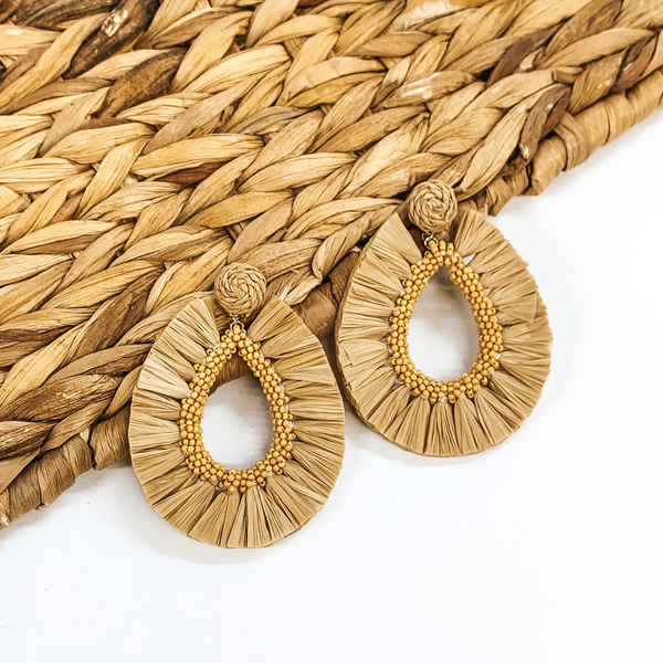 Circle beaded earrings with a hanging open teardrop pendant. The pendant is a beaded teardrop with raffia wrapped around the edges. The earrings are tan colored. These earrings are pictured on a white backround while partially laying on a tan woven material.