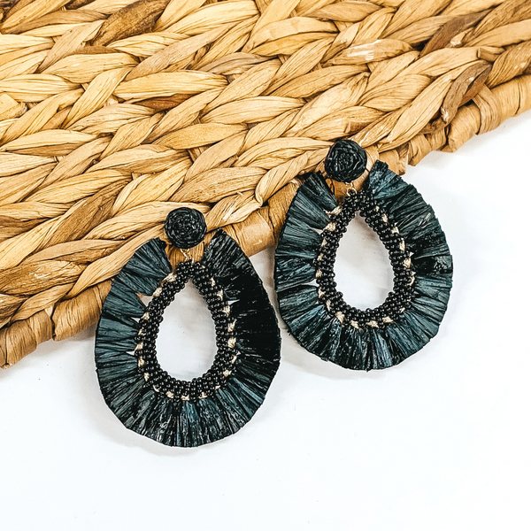 Circle beaded earrings with a hanging open teardrop pendant. The pendant is a beaded teardrop with raffia wrapped around the edges. The earrings are black colored. These earrings are pictured on a white backround while partially laying on a tan woven material. 