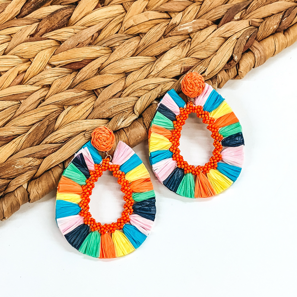 Circle beaded earrings with a hanging open teardrop pendant. The pendant is a beaded teardrop with raffia wrapped around the edges. The earrings are multicolored. These earrings are pictured on a white backround while partially laying on a tan woven material.  