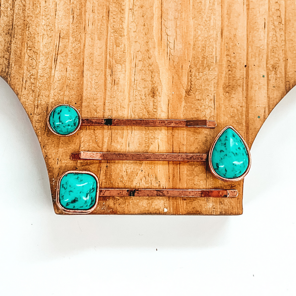 Set of three copper hair pins. Each hair pin has a single turquoise stone in a different shape. These shapes include a circle, teardrop, and a square. These hair pins are pictured on a brown block on a white background. 