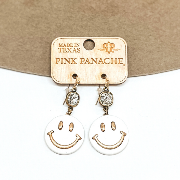 Bronze fish hook earrings with a clear cushion cut crystal in a bronze setting. Hanging from the bottom of the crystal is a white circle pendant with a gold smiley face on the front. These earrings are pictured on a brown and white background. 